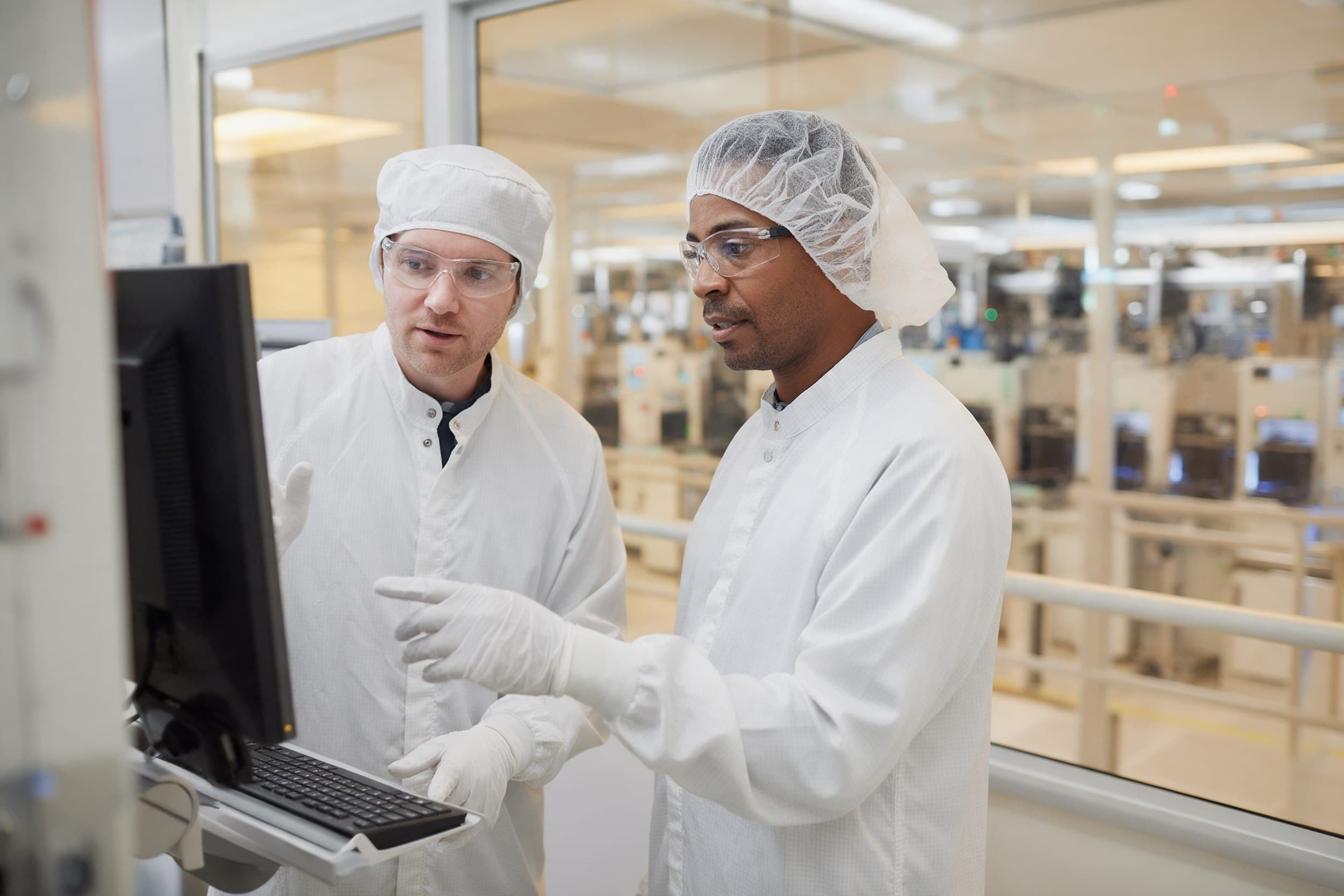 Two engineers in cleanroom gowning reviewing tool data on a screen at a fab mezzanine