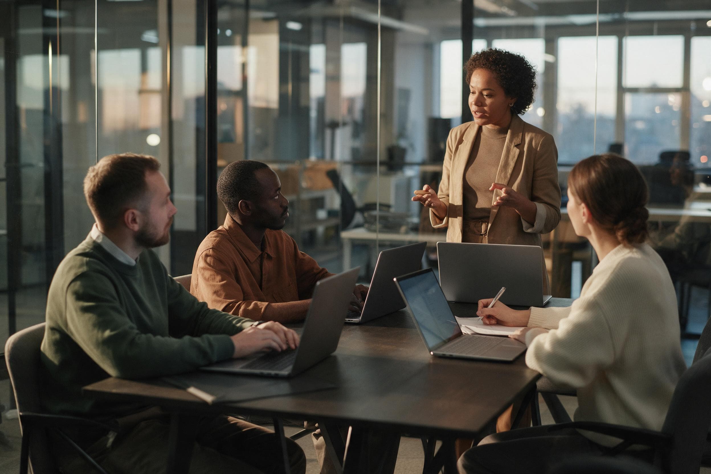 A contract specialist standing at the end of a meeting room table explaining something to a client team over open laptops